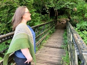 Louise is standing on a footbridge in a wood. She is wearing a blue handmade top and she holds a triangular shape garter stitch shawl