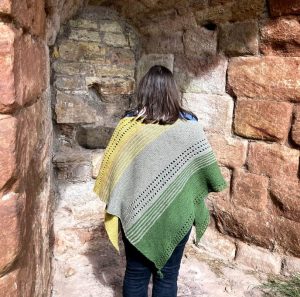 Louise stands facing away from the camera, she is wearing a triangular shawl in yellow, green and mushroom colours.