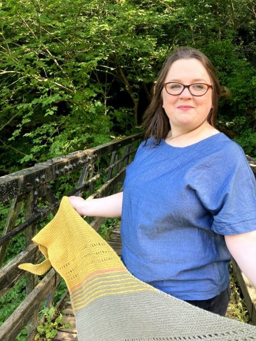 Louise is standing on a footbridge in a wood. She is wearing a blue handmade top and she holds a triangular shape garter stitch shawl