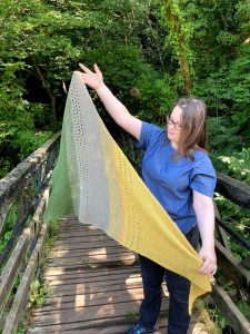 Louise is standing on a footbridge in a wood. She is wearing a blue handmade top and she holds a triangular shape garter stitch shawl